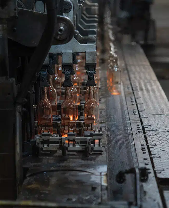 Rows of newly formed clear glass bottles being handled by automated machinery on a production line, with orange light glowing through the glass inside the industrial manufacturing equipment.