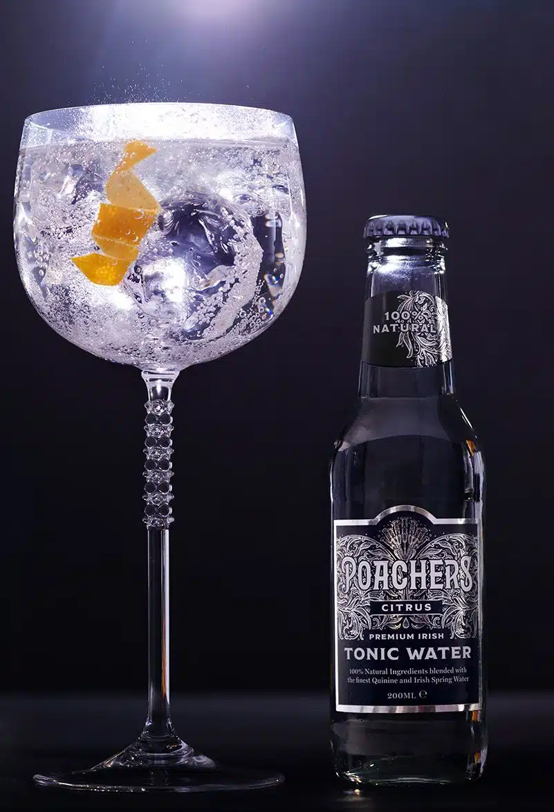 Bottle of Poachers Citrus Premium Irish Tonic Water beside a large gin glass filled with ice, tonic and an orange twist, set against a dark studio background with dramatic lighting.