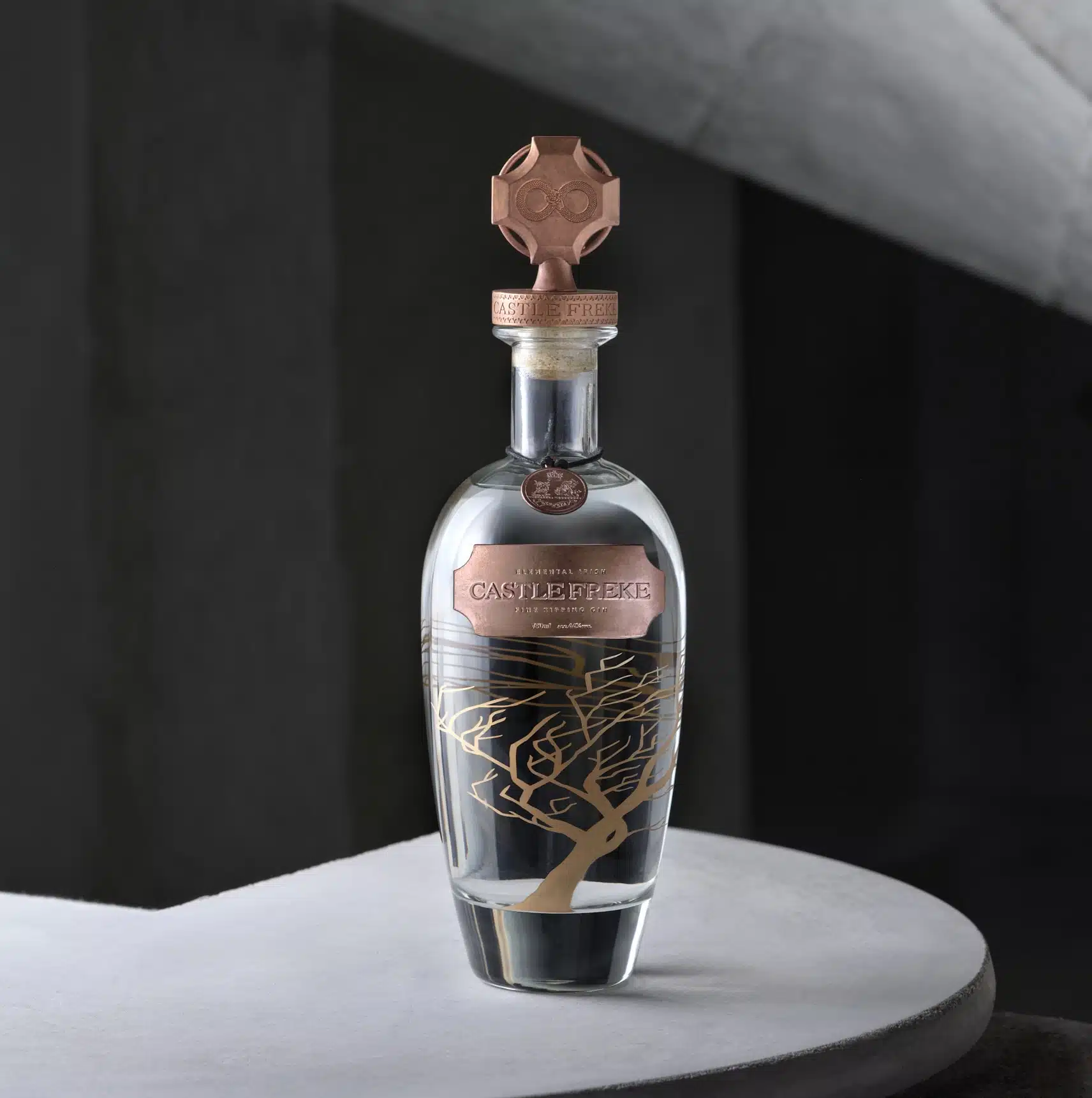 Bottle of Castle Freke Irish Spirit displayed on a curved stone surface, featuring a clear glass bottle with a copper-toned label, decorative tree illustration, and ornate cross-shaped stopper. The product is photographed against a dark, architectural background with soft lighting that highlights the bottle’s premium design.