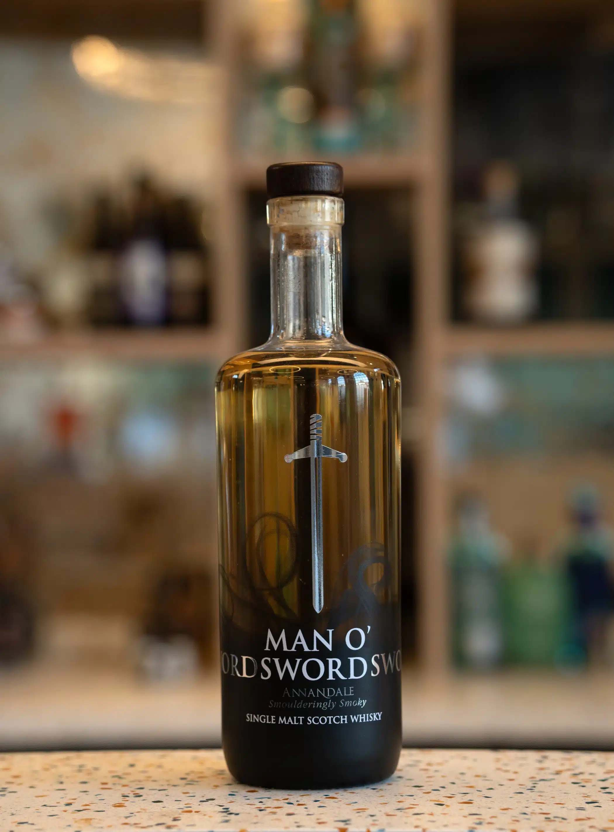 Bottle of Man O’ Swords single malt Scotch whisky by Annandale Distillery on a speckled countertop, featuring a dark label with a silver sword illustration and amber whisky visible through the glass. The bottle is photographed against softly blurred shelves of spirits in a warm, atmospheric setting.