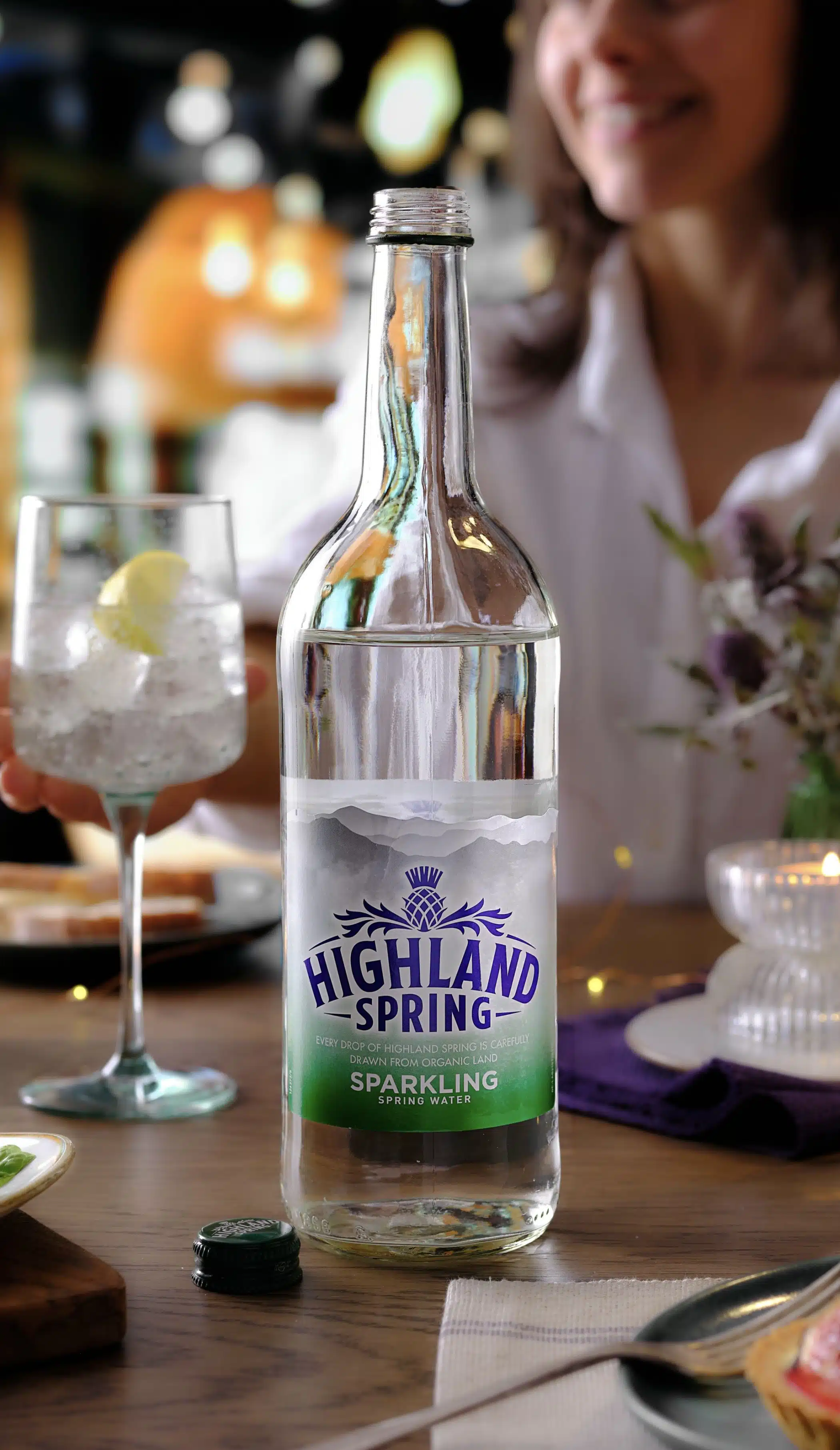 Highland Spring still and sparkling water bottles on a restaurant table with food, ice-filled glasses and soft ambient lighting in the background.