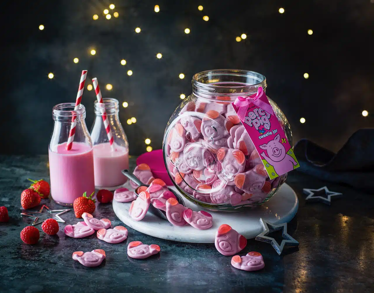 A glass jar filled with Percy Pig gummy sweets, surrounded by scattered sweets, strawberries, and raspberries. Two small glass bottles with pink milk and striped straws are in the background, along with twinkling lights