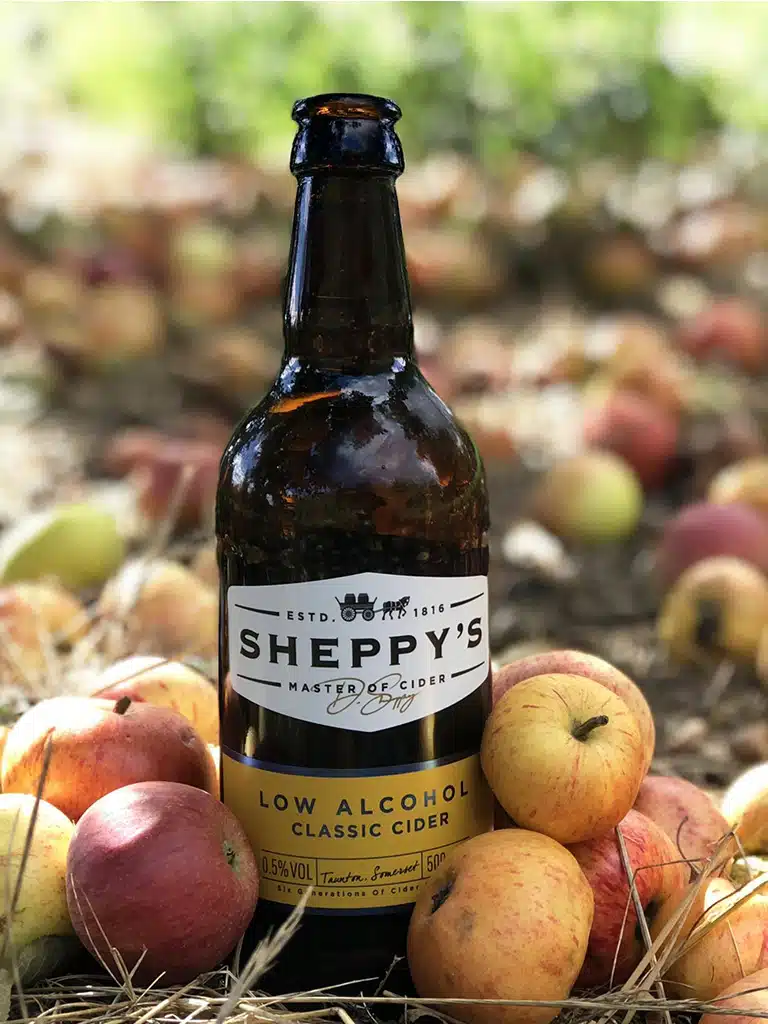 Amber glass bottle of Sheppy's Low Alcohol Classic Cider standing amongst freshly fallen apples in an orchard, showcasing the traditional 500ml AMC cider bottle shape.