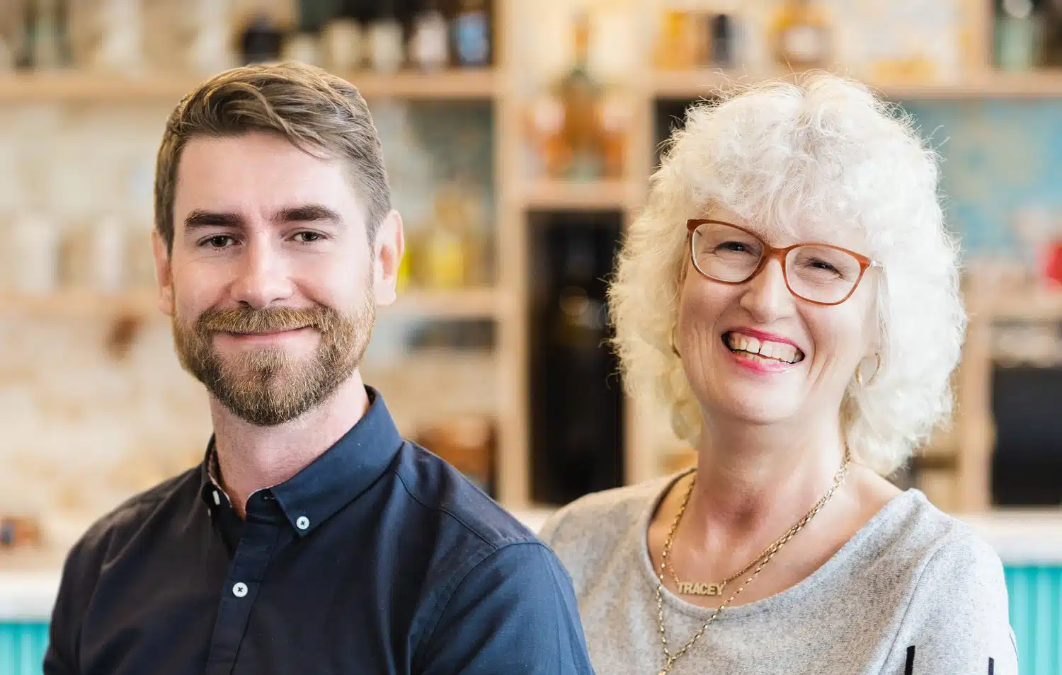 2 staff members sat in front of a bar. Man, left, in blue shirt, woman, right, in grey top.