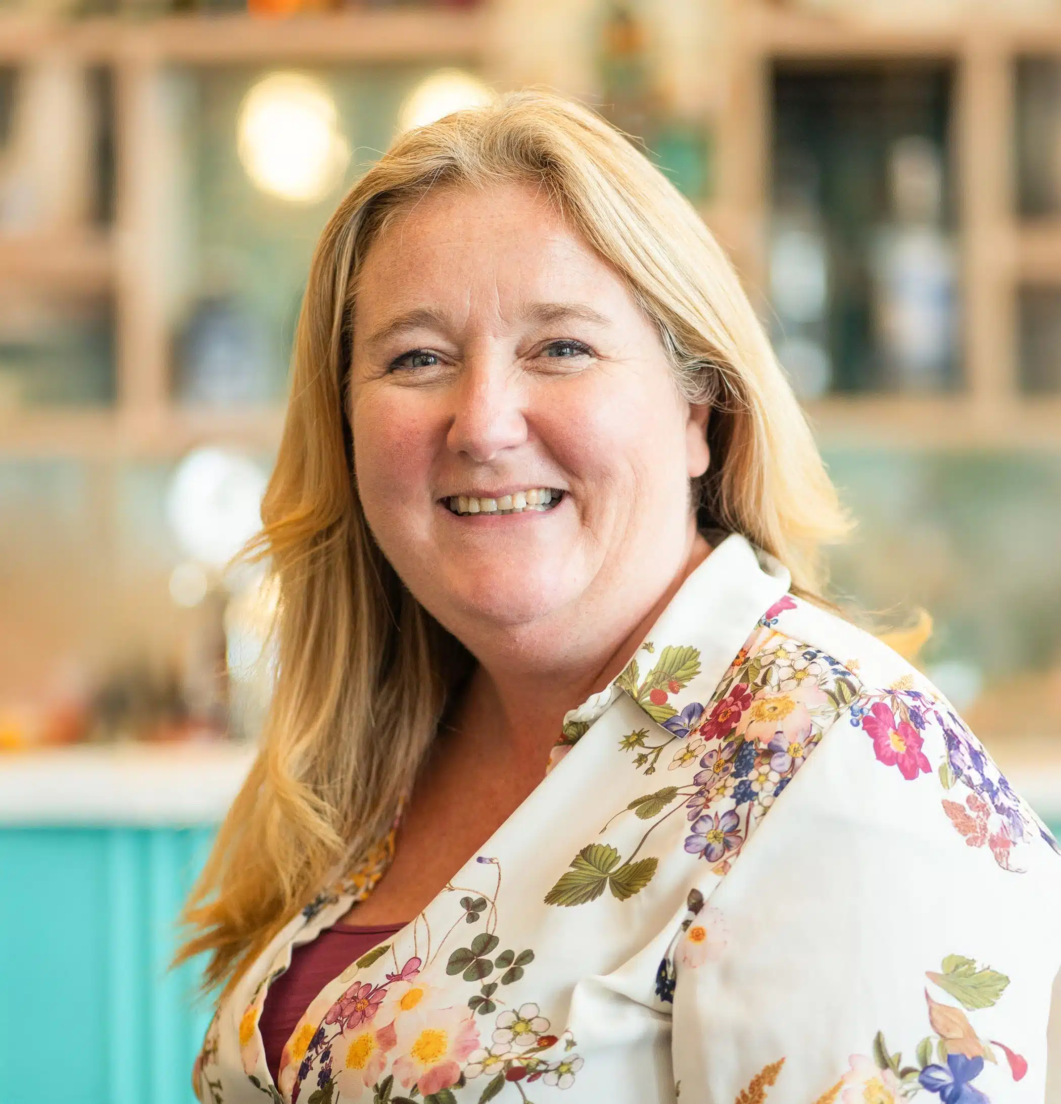 A smiling woman with long blonde hair wearing a cream blouse decorated with colorful floral patterns, photographed indoors with a softly blurred background.