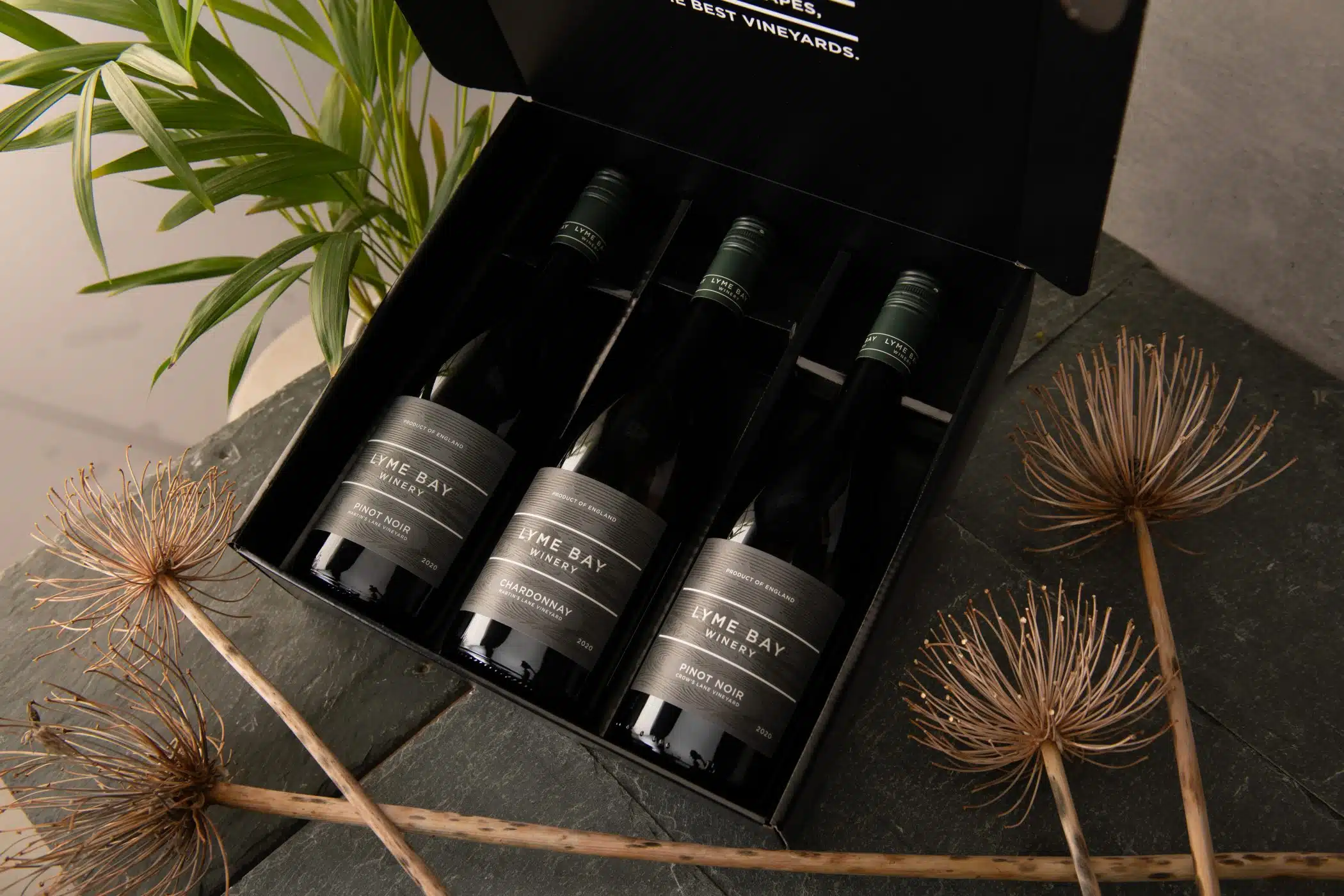 Gift box containing three bottles of Lyme Bay Winery wine — Pinot Noir, Chardonnay, and Pinot Noir — displayed on a stone surface with dried flowers and green foliage.