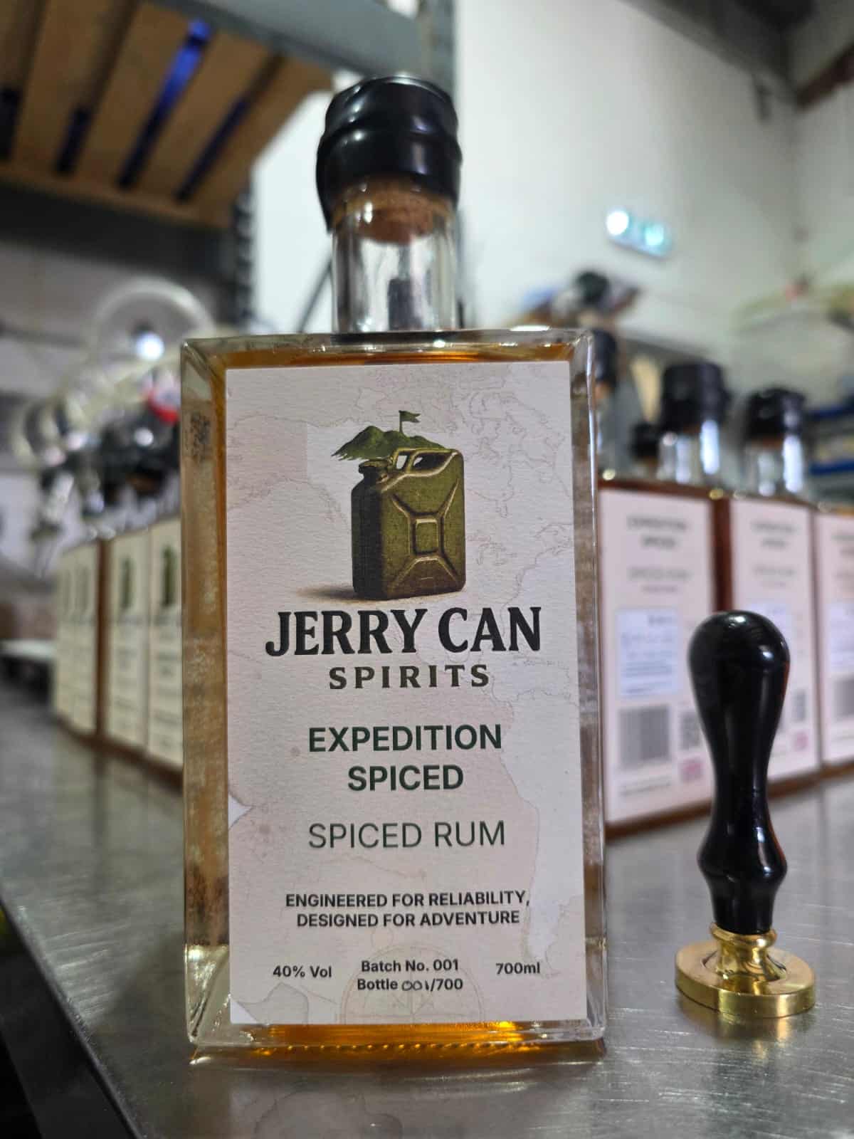 Square glass bottle of Jerry Can Spirits Expedition Spiced Rum with a natural cork sealed in black wax, displayed on a stainless steel workbench beside a wax seal stamp.