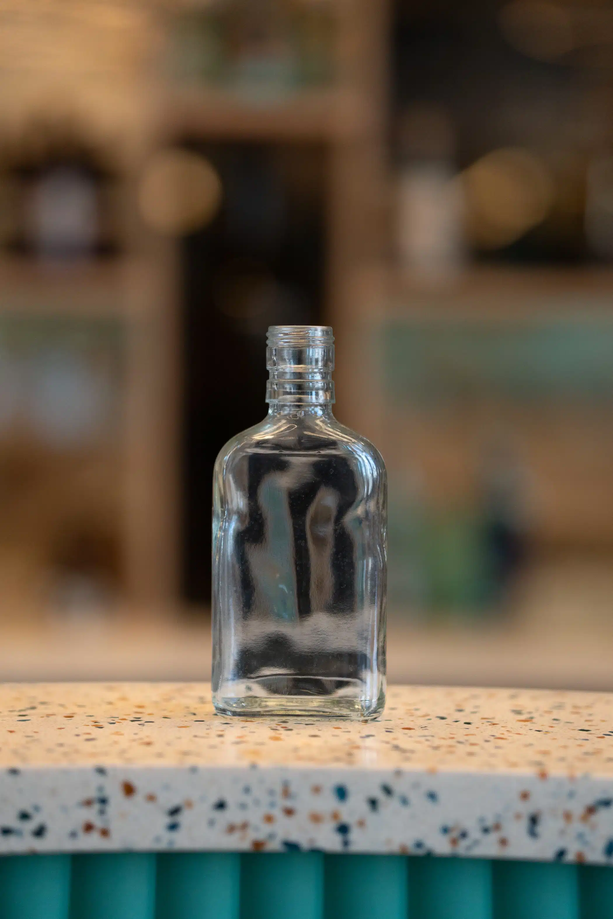 Clear glass bottle with a rectangular body, rounded shoulders and threaded screw-top neck, displayed on a terrazzo-style countertop with softly blurred shelving in the background.